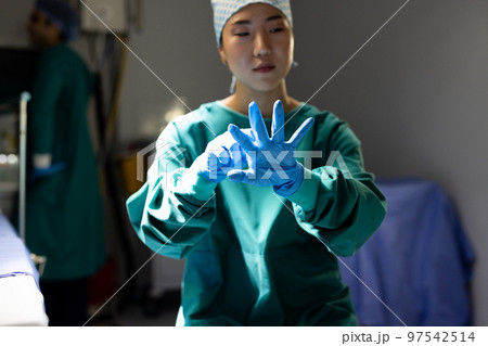 Asian female surgeon in surgical cap and gown putting on gloves in operating theatre 97542514