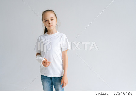 Portrait of sad blonde little girl with broken hand wrapped in white plaster bandage standing on white isolated background in studio, looking at camera. Concept of child insurance and healthcare. 97546436