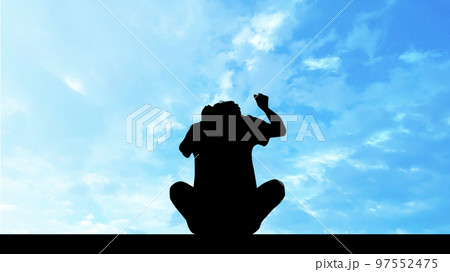 地面に座って腕を振り上げている男性シルエット_青空背景_ワイド 97552475
