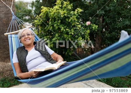 Smiling senior good-looking grey hair woman wearing glasses while reading in hammock in the summer garden Smiling senior good-looking grey hair woman wearing glasses while reading in hammock in the summer garden 97553869