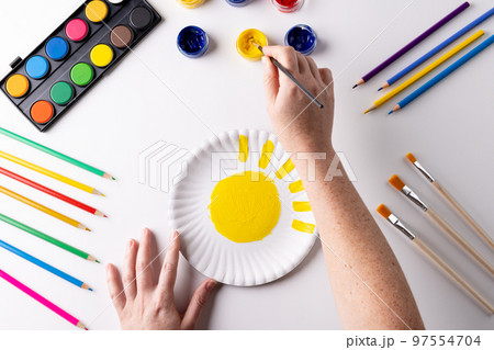 Overhead of hands painting with yellow paint on paper plate, with art materials on table top 97554704