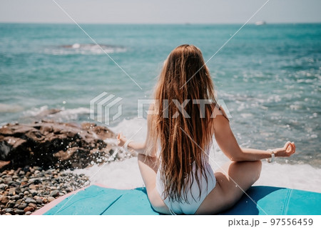 Woman yoga sea. Young woman with long hair in white swimsuit and boho style braclets practicing outdoors on yoga mat by the ocean on sunny day. Women's yoga fitness routine. Healthy lifestyle, harmony Woman yoga sea. Young woman with long hair in white swimsuit and boho style braclets practicing outdoors on yoga mat by the ocean on sunny day. Women's yoga fitness routine. Healthy lifestyle, harmony 97556459