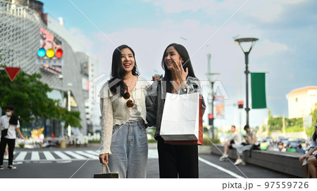 Fashionable women friends with shopping bags standing in city avenue with blurred cityscape and blue sky in background Fashionable women friends with shopping bags standing in city avenue with blurred cityscape and blue sky in background 97559726