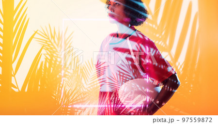 Composite of biracial female rugby player with ball standing over illuminated square and plants 97559872