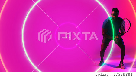 African american male player holding racket and standing over illuminated circles on pink background 97559881