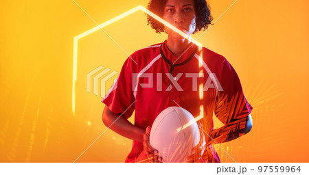 Confident biracial female player with ball and illuminated hexagon and plants on yellow background 97559964