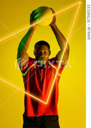African american male rugby player throwing ball over illuminated triangle on yellow background 97560232