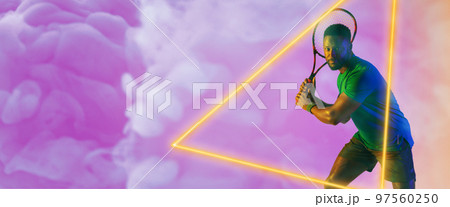 African american male tennis player holding racket by illuminated triangle over smoky background African american male tennis player holding racket by illuminated triangle over smoky background 97560250