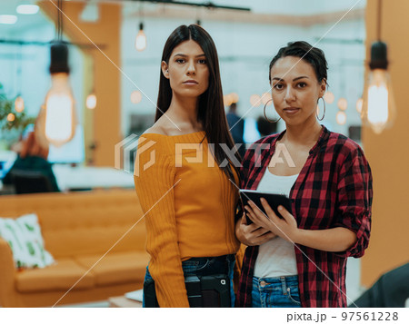 Portrait of two businesswomen talking to each other while standing in a modern business office with their colleagues using laptop and tablet.. Marketing concept. Multi-ethnic society. Portrait of two businesswomen talking to each other while standing in a modern business office with their colleagues using laptop and tablet.. Marketing concept. Multi-ethnic society. 97561228