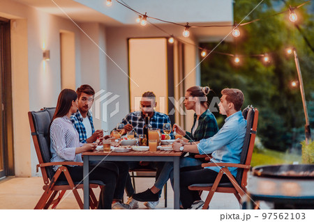 A group of young diverse people having dinner on the terrace of a modern house in the evening. Fun for friends and family. Celebration of holidays, weddings with barbecue. 97562103