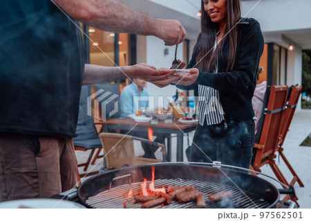 A group of friends and family barbecue together in the evening on the terrace in front of a large modern house A group of friends and family barbecue together in the evening on the terrace in front of a large modern house 97562501