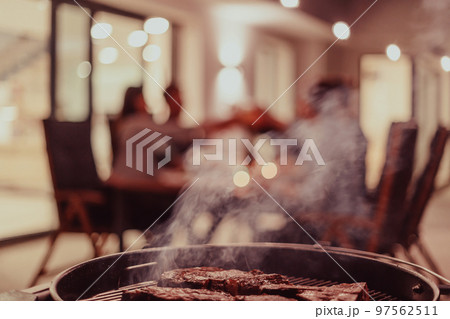 Close-up photo of delicious meat being grilled. In the background, friends and family are sitting and waiting for a meal 97562511
