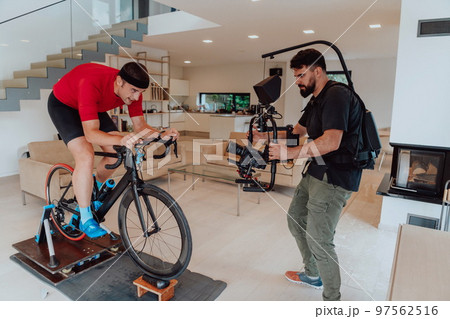 A cameraman filming an athlete riding a triathlon bike on a simulation machine in a modern living room. Training in pandemic conditions. A cameraman filming an athlete riding a triathlon bike on a simulation machine in a modern living room. Training in pandemic conditions. 97562516