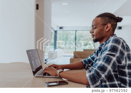 African American man in glasses sitting at a table in a modern living room, using a laptop for business video chat, conversation with friends and entertainment African American man in glasses sitting at a table in a modern living room, using a laptop for business video chat, conversation with friends and entertainment 97562592