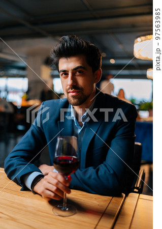 Vertical portrait of confident elegant man in fashion suit holding glasses of red wine sitting at table in restaurant with dark interior, looking at camera. Bearded male resting, having dinner alone. Vertical portrait of confident elegant man in fashion suit holding glasses of red wine sitting at table in restaurant with dark interior, looking at camera. Bearded male resting, having dinner alone. 97563985