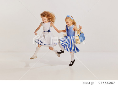 Portrait of two little girls, children in cute dresses posing, running, jumping over grey background. Playful kids 97566067
