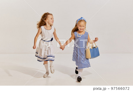 Portrait of two little girls, children in cute dresses posing, smiling, holding hands and walking over grey background 97566072