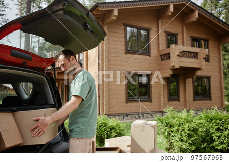 Man unloading boxes from car trunk while moving into new wooden house in nature 97567963
