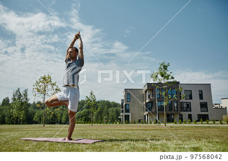 Young man doing yoga outdoors on lawn in modern housing complex 97568042