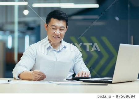 Portrait of a young Asian male accountant sitting at a desk in the office, holding documents in his hands, using a calculator. Works with accounts, budget, loans. 97569694