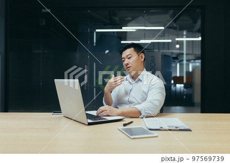 A young Asian man is sitting in the office at the table, working on a laptop. He waves his hand in his face. He feels bad, feels hot, needs fresh air, medical help. A young Asian man is sitting in the office at the table, working on a laptop. He waves his hand in his face. He feels bad, feels hot, needs fresh air, medical help. 97569739