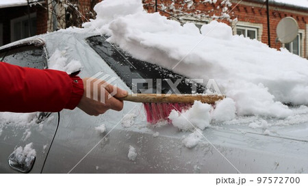 a man in a red jacket brushes a gray car from a large amount of fresh snow after a snow storm, cleaning the back of the car from a high layer of snow after a snowfall 97572700