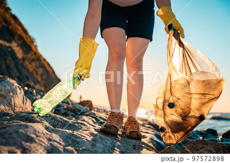 Person volunteer collects garbage in a plastic bag. In the background the sea and the sunset. Close up of woman's legs. Bottom view. The concept of coastal zone cleaning and Earth Day 97572898