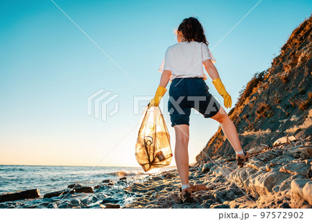 A female volunteer stands with a plastic bag in her hand. Back view. Cleaning of the coastal zone and wild beaches. The concept of Earth Day 97572902