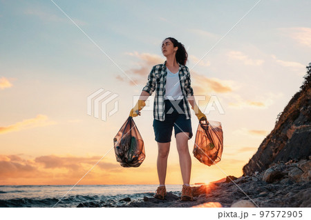 A female volunteer carries two large bags of garbage in her hands. In the background, the sea and the sunset. Copy space. The concept of environmental protection and coastal zone cleaning 97572905