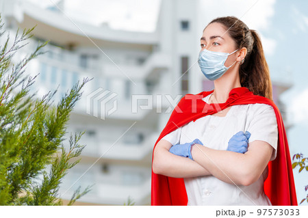 A young female doctor wearing a surgical mask and rubber gloves poses with her arms crossed and a super hero's cape on her back. The hospital is blurred in the background. The concept of hero doctors A young female doctor wearing a surgical mask and rubber gloves poses with her arms crossed and a super hero's cape on her back. The hospital is blurred in the background. The concept of hero doctors 97573033