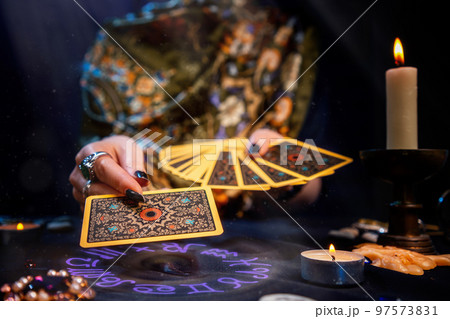 The fortune teller holds a fan of cards in her hands and holds out a card with one hand. The astrological circle glows at hand. Close-up. The concept of divination, magic and esotericism The fortune teller holds a fan of cards in her hands and holds out a card with one hand. The astrological circle glows at hand. Close-up. The concept of divination, magic and esotericism 97573831
