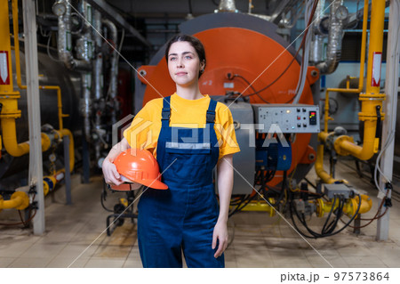 The concept of industrial manufactory. A young smiling woman in uniform holds a safety helmet in her hand and poses while standing in the boiler room 97573864
