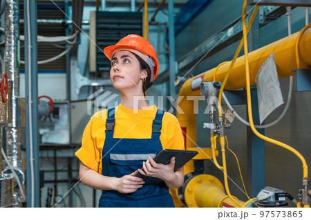 Production inspection. A young engineer in a uniform and a protective helmet, holding a tablet in his hands and conducting an inspection of the equipment. The concept of human equality Production inspection. A young engineer in a uniform and a protective helmet, holding a tablet in his hands and conducting an inspection of the equipment. The concept of human equality 97573865