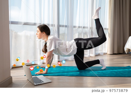 Young caucasian woman in sportswear doing yoga live online. Indoor. Emoticons and emojis fly around the laptop. The concept of social networks, online training and blogging Young caucasian woman in sportswear doing yoga live online. Indoor. Emoticons and emojis fly around the laptop. The concept of social networks, online training and blogging 97573879