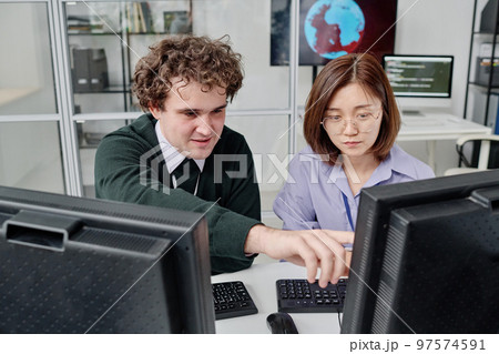 Two young colleagues working with security of computer program together, they sitting at table with computer monitors Two young colleagues working with security of computer program together, they sitting at table with computer monitors 97574591
