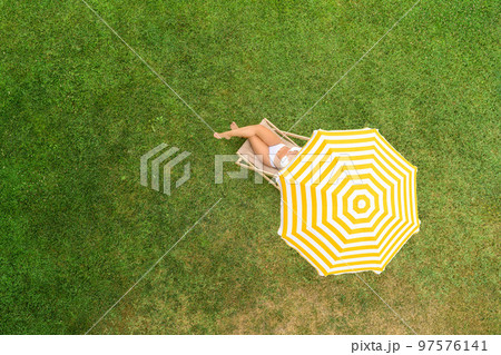 Woman in a white bikini sitting on deck chair under yellow umbrella on the green grass sunbathes at summer day. Top view, drone, aerial view, Woman in a white bikini sitting on deck chair under yellow umbrella on the green grass sunbathes at summer day. Top view, drone, aerial view, 97576141