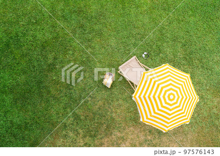 Yellow umbrella with deck chair, basket for picnic, headphones on the green grass at summer day. Top view, drone, aerial view. Yellow umbrella with deck chair, basket for picnic, headphones on the green grass at summer day. Top view, drone, aerial view. 97576143