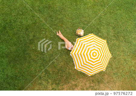 Woman in white bikini with basket for picnic lying under yellow umbrella on the green grass sunbathes at summer day. Top view, drone, aerial view. Woman in white bikini with basket for picnic lying under yellow umbrella on the green grass sunbathes at summer day. Top view, drone, aerial view. 97576272