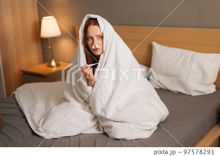 Portrait of sick sad woman suffering from runny nose disease, paper tissues inserted into nose, measuring temperature with thermometer sitting in bed covered with warm blanket at home. 97578291
