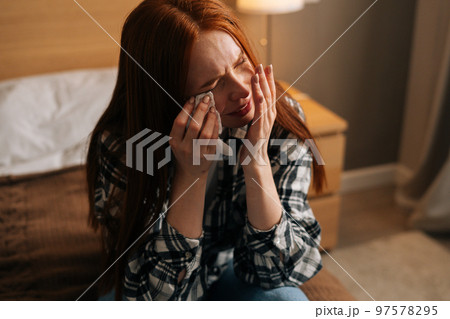Close-up portrait of unhappy depressed redhead woman crying and wiping tears with paper tissue, suffer from grief, divorce or break up with boyfriend, broken heart, sitting on bed at home. 97578295