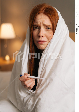 Vertical portrait of ill exhausted woman suffering from runny nose disease with paper tissues into nose, measuring temperature with thermometer, sitting in bed covered with blanket, looking at camera. 97578296