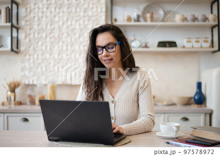 Happy young lady freelancer working from home, sitting at table and using laptop computer, kitchen interior 97578972