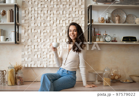Carefree happy lady relaxing in cozy kitchen, sitting at counter and drinking coffee, smiling at camera, copy space 97579022