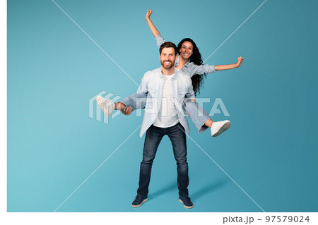 Cheerful young european guy hold woman in casual on back, have fun, isolated on blue background, studio 97579024