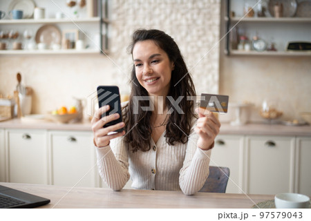 Happy caucasian woman buying food online, using cellphone and plastic credit card, shopping online, sitting in kitchen 97579043