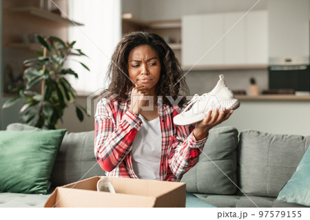Pensive millennial black lady unpacks cardboard box, holds shoes, thinks in room interior 97579155