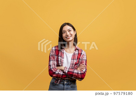 Dreamy positive asian lady standing with folded arms and looking aside, posing over yellow background, copy space 97579180