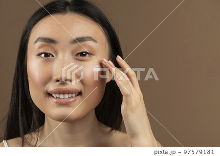 Portrait of asian lady applying anti-aging or moisturising face cream, posing over brown studio background, copy space 97579181