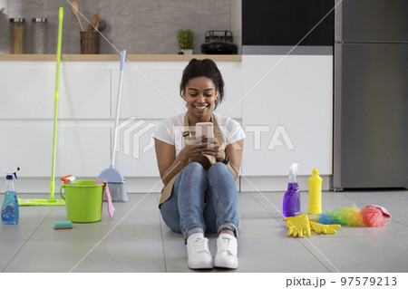 Happy millennial african american lady in apron sitting on floor with cleaning supplies typing on smartphone Happy millennial african american lady in apron sitting on floor with cleaning supplies typing on smartphone 97579213