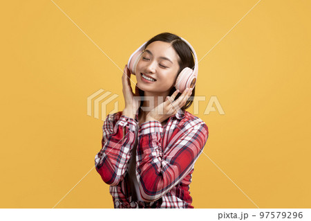 Peaceful asian woman listening to music online in wireless headphones and touching new headset, yellow background Peaceful asian woman listening to music online in wireless headphones and touching new headset, yellow background 97579296